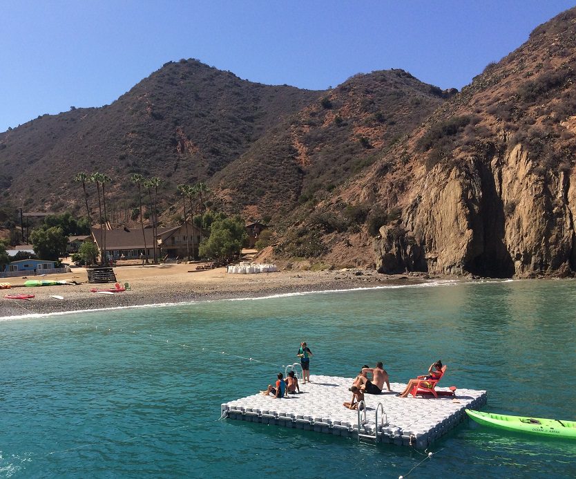 A family sits on a floating dock that is off shore. They are in a tropical mountainous area.