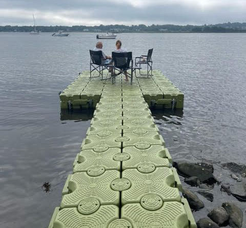 A couple sits in chairs on a green floating dock, looking across the lake.